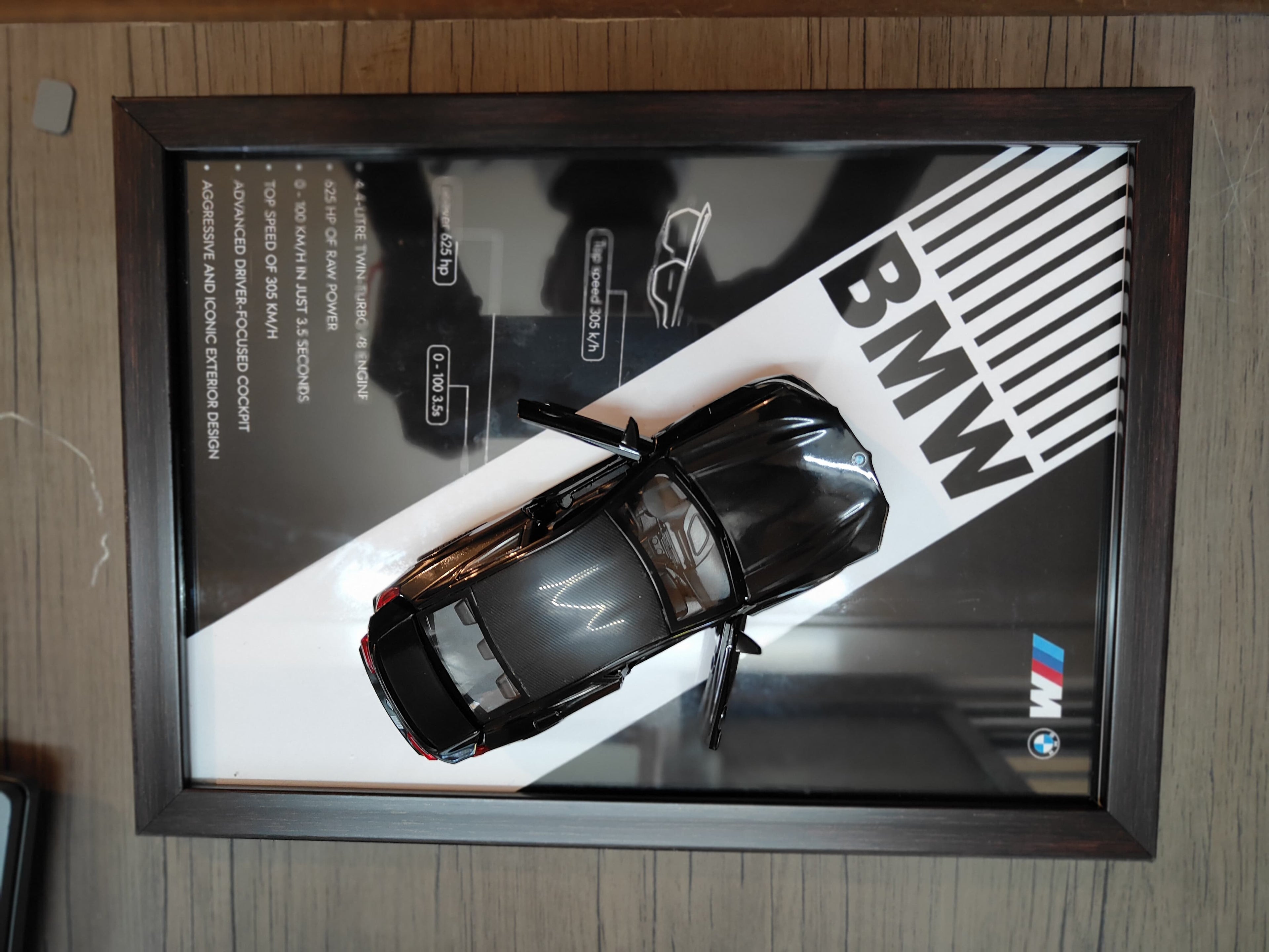 BMW M Series gloss black die-cast model in dark wood shadow box with BMW M branded backdrop and performance specs